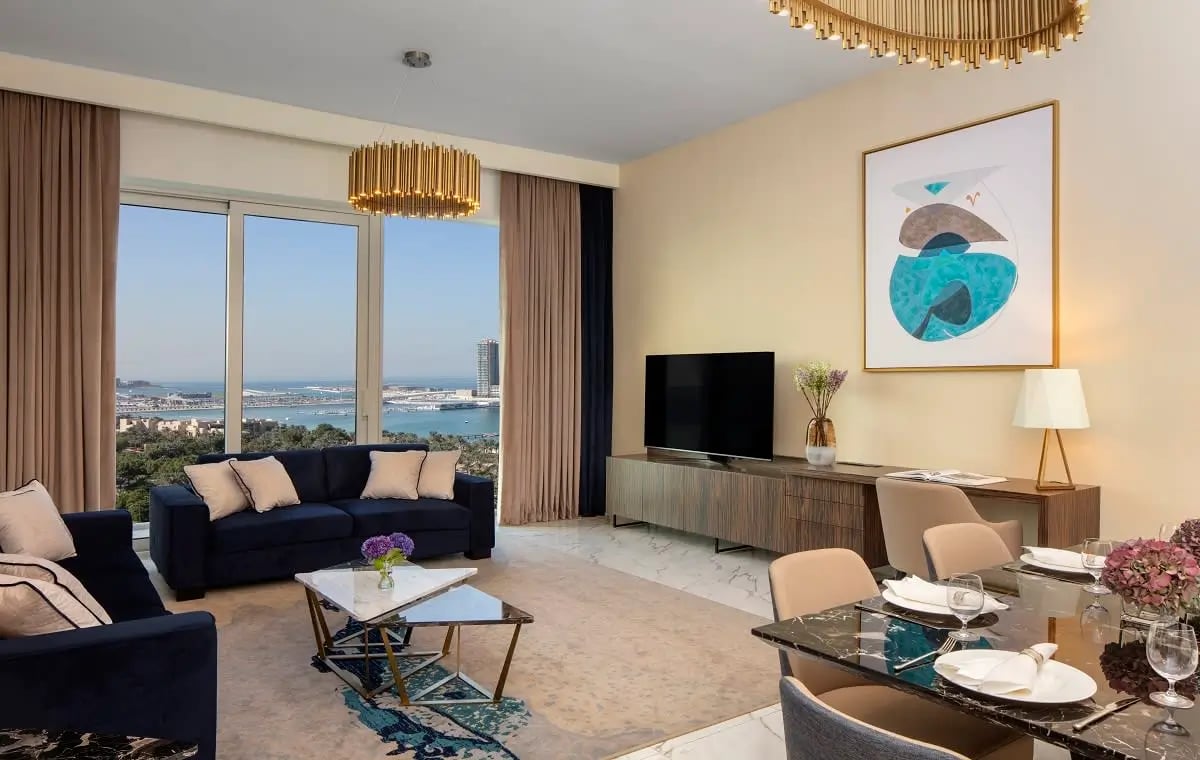 Luxury modern hotel living room with elegant navy blue sofas, marble coffee table, gold chandeliers, and a stunning sea view.