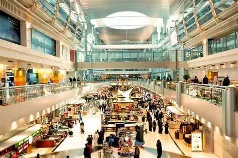 Spacious and brightly lit airport duty-free shopping area with multiple retail stores, kiosks, and shoppers. The scene features high ceilings, modern architecture, and a bustling atmosphere.