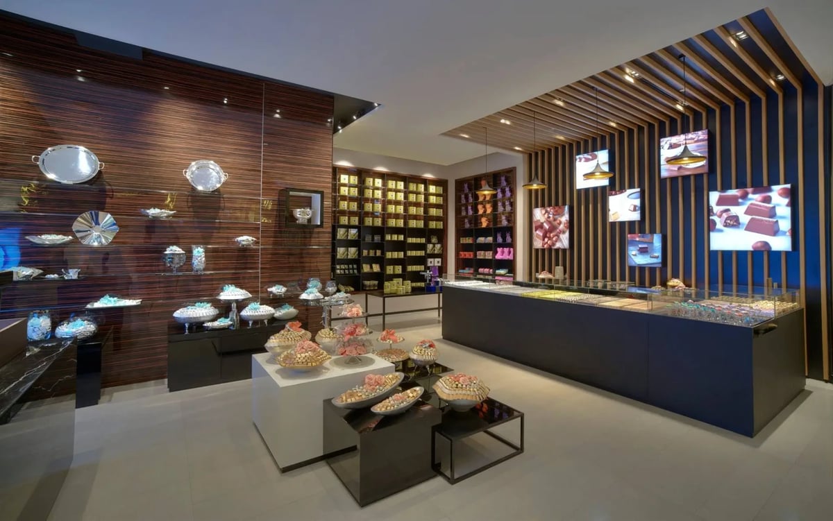 Modern luxury chocolate boutique interior featuring dark wood walls, illuminated display shelves with assorted chocolates, elegant trays on tables, and a sleek glass counter. The space is accented with stylish lighting and digital screens displaying chocolate imagery.