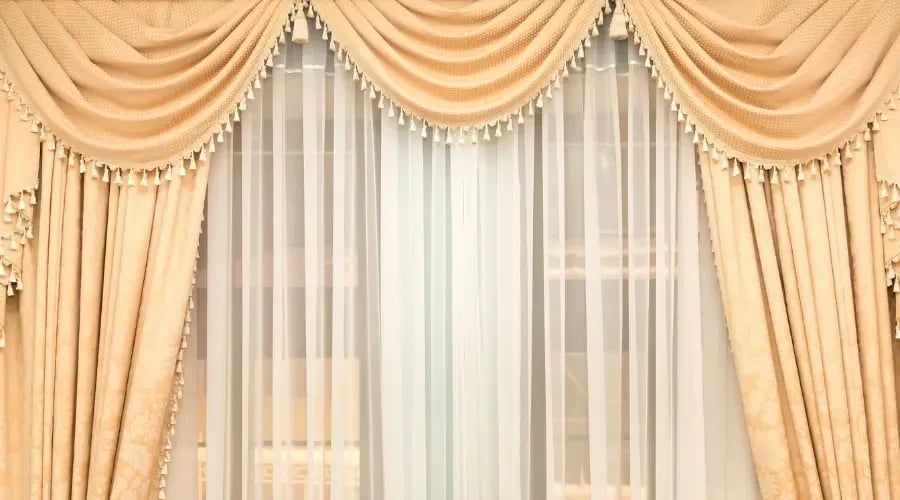 Elegant cream-colored curtains with a draped swag design, adorned with tassels, paired with sheer white curtains underneath for a luxurious, classic look.
