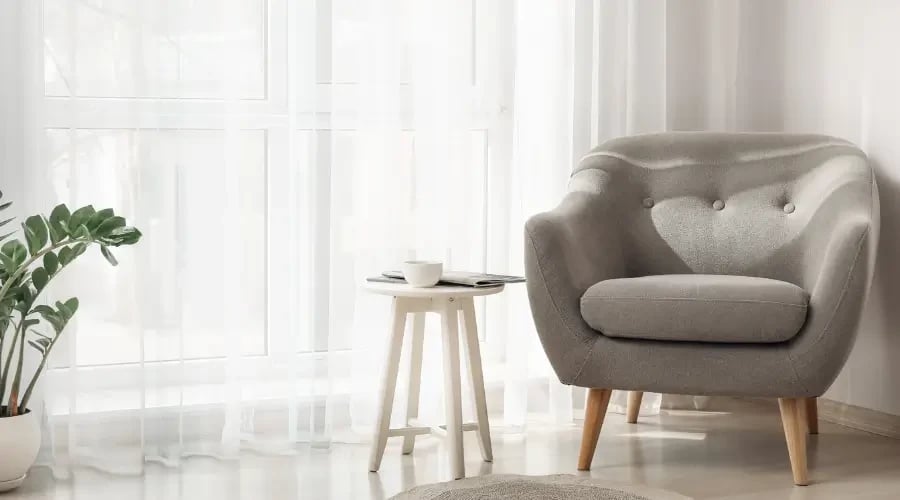 Minimalist corner with a soft gray armchair, a small side table, and sheer white curtains, allowing natural light to flood the space. A green plant adds a touch of freshness to the serene, calm environment.