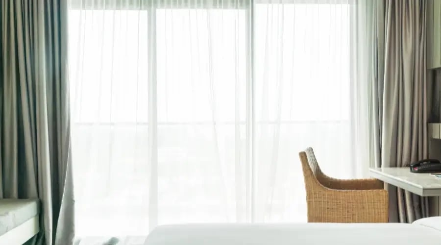 Bright bedroom with sheer and layered curtains, allowing natural light to filter through while providing privacy, complemented by a simple wicker chair and minimalist decor.