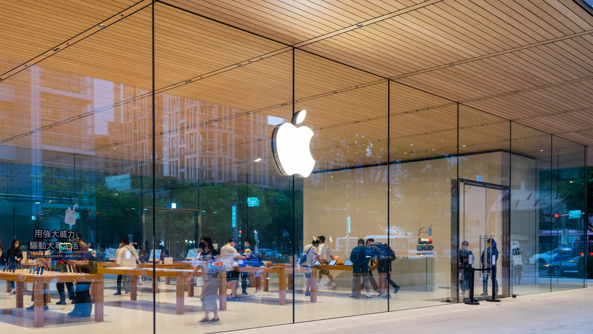Apple retail store exterior with large glass windows, wooden ceiling, and illuminated Apple logo. Shoppers are visible inside the modern, minimalist store interior.