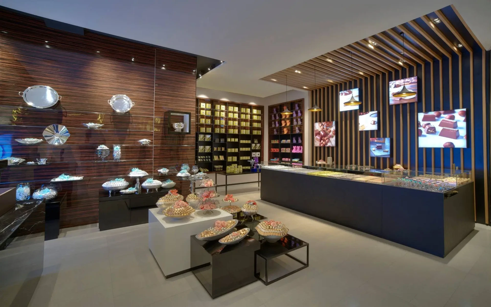 Modern luxury chocolate boutique interior featuring dark wood walls, illuminated display shelves with assorted chocolates, elegant trays on tables, and a sleek glass counter. The space is accented with stylish lighting and digital screens displaying chocolate imagery.