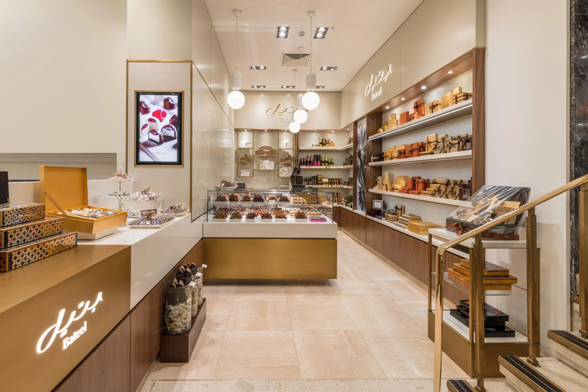 Luxury chocolate boutique interior with elegant display cases, gold and wood shelving, and a variety of boxed chocolates and sweets. The space features warm lighting, modern design elements, and the Bateel logo illuminated on the counter.