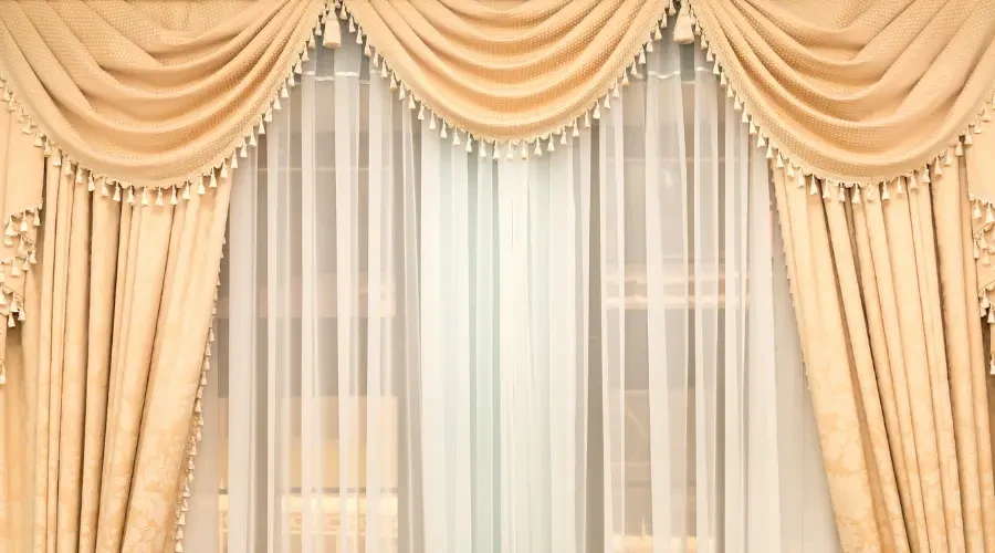 Elegant cream-colored curtains with a draped swag design, adorned with tassels, paired with sheer white curtains underneath for a luxurious, classic look.