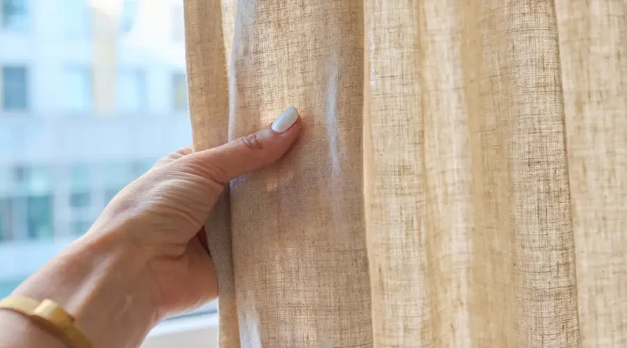 Close-up of a hand gently pulling back a natural linen curtain, showcasing the soft, textured fabric that blends seamlessly with minimalist interior design, highlighting the natural material.