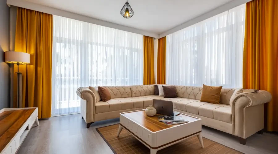 Bright living room with vibrant yellow curtains paired with sheer white curtains, creating a lively and airy atmosphere. The space features a beige sofa, a coffee table with a laptop, and modern lighting.